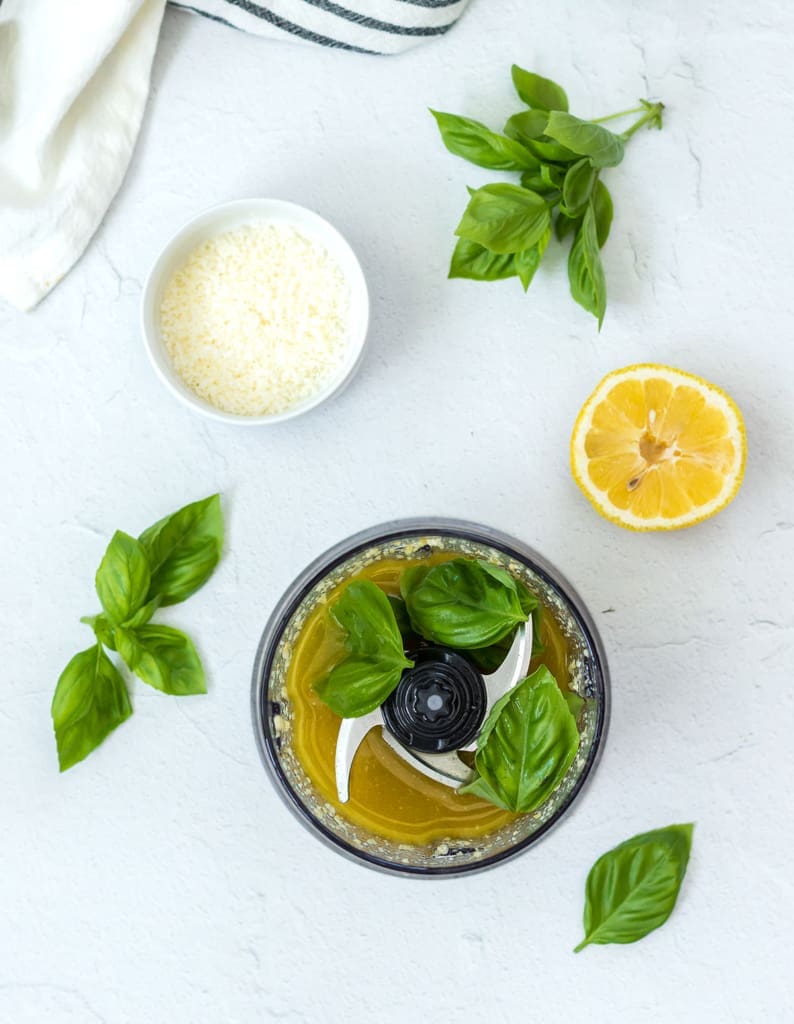 Basil and olive oil in a blender next to basil leaves and lemon.