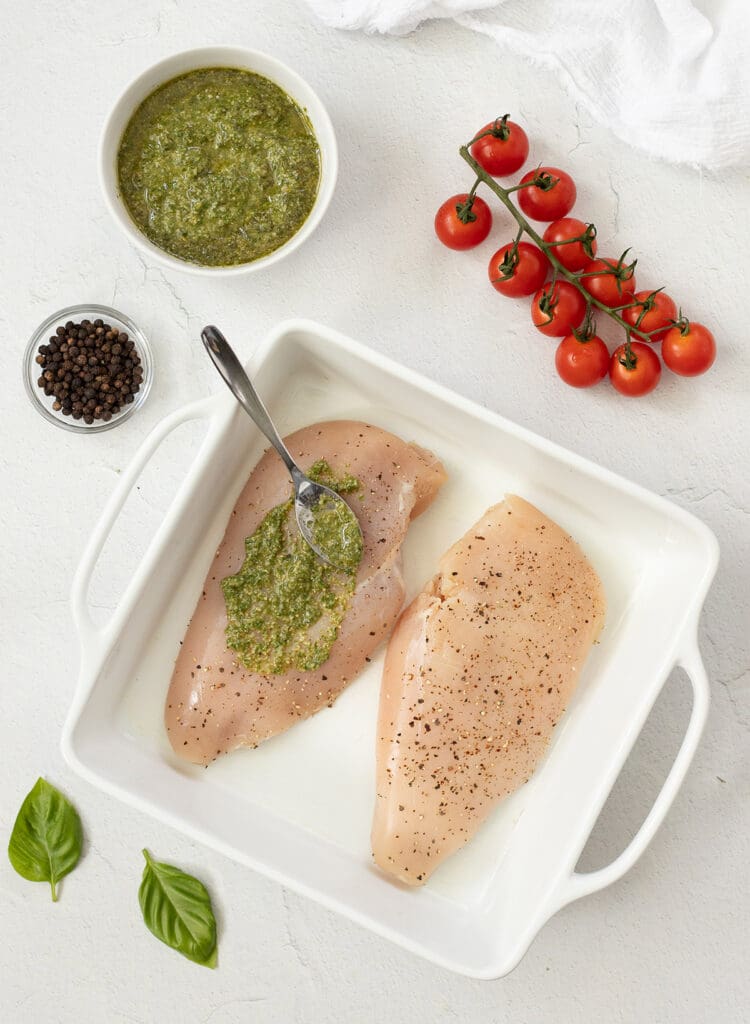 Spoon of pesto being rubbed on a raw chicken breast.